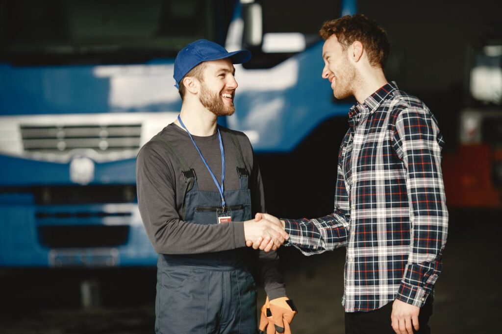 Two men shaking hands and smiling in front of a blue semi-truck, one wearing a cap and overalls with a lanyard, the other in a plaid shirt.