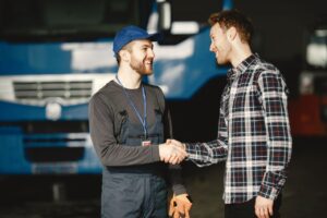 Two men shaking hands and smiling in front of a blue semi-truck, one wearing a cap and overalls with a lanyard, the other in a plaid shirt.