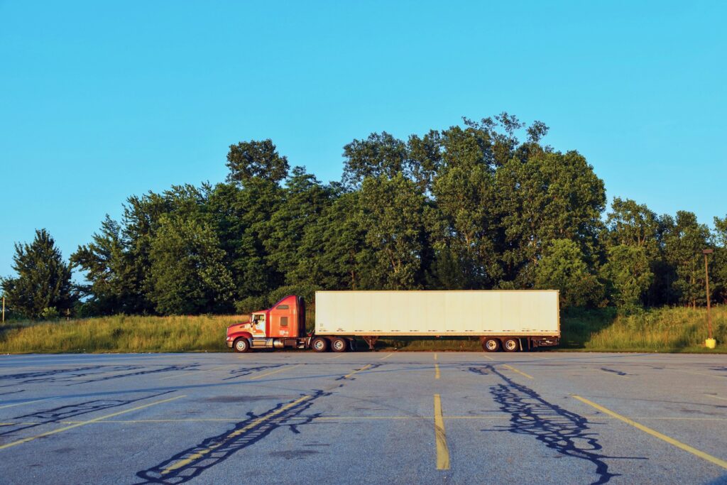 Red semi-truck with a white cargo trailer parked in an empty lot at golden hour, representing the older commercial trucks owner-operators often finance