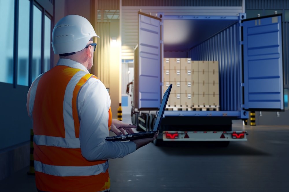 Logistic work. Container truck. Man logistician with his back to camera. Unloading truck in warehouse. Logistician with laptop. Man near industrial buildings. Truck with boxes on pallets