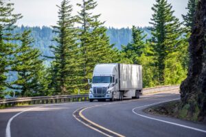 Big rig white powerful American bonnet long haul semi truck transporting commercial cargo in refrigerated semi trailer moving uphill on winding road with a green trees and safety fence on the side