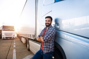Portrait of young Caucasian bearded trucker with arms crossed standing by his truck vehicle.