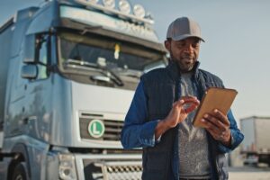 African American professional truck driver setting up navigation for destination. Checking his route on tablet computer and standing by long vehicle.