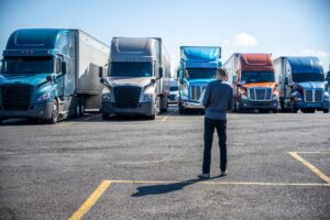 Slim Truck driver carries a box with purchases in his hands and going to his big rig semi ruck parked on the truck stop parking lot standing in row with another semi trucks with semi trailers
