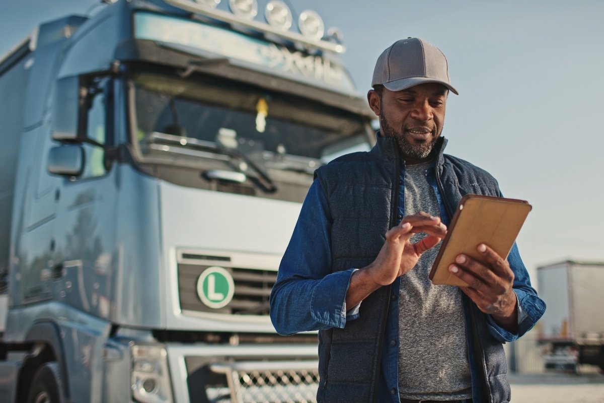African American professional truck driver setting up navigation for destination. Checking his route on tablet computer and standing by long vehicle. Transportation service.