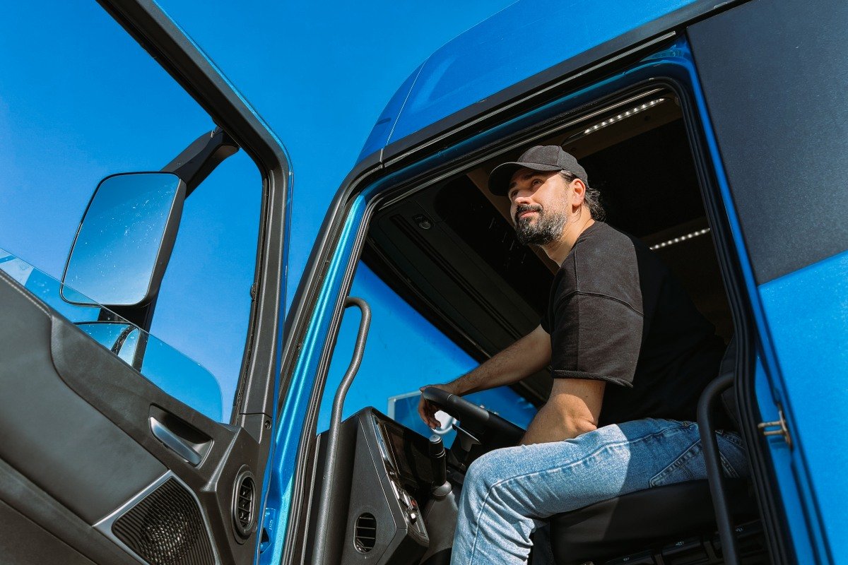 A young man is a professional driver in the cab of a blue truck. Handsome Caucasian guy, a transportation worker sits in a big truck car