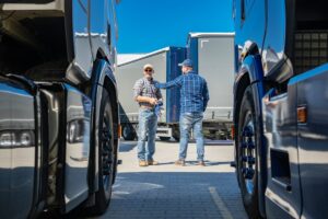 Two truck drivers engage in a conversation while standing between parked trucks in a transport yard.