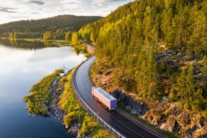 Aerial view of semi truck with cargo trailer on road curve at lake shore with green pine forest.