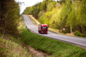 Red truck with tarpaulin semitrailer transports cargo on country road.