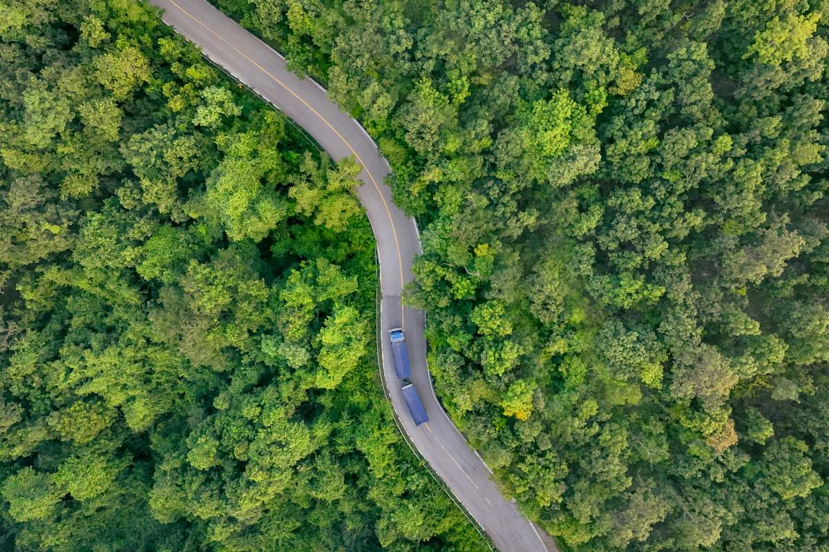 Aerial view hydrogen truck on forest highway. Sustainable transportation. Blue zero emission vehicle driving through green landscape. Eco-friendly logistics solution. Clean energy freight delivery.