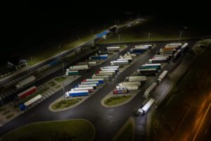 nighttime aerial shot shows a well-lit truck stop where many semi-trucks are parked in neat rows.
