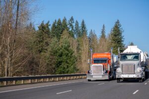 Two different big rig orange and white semi trucks with tank semi trailers transporting liquid or liquefied chemicals commercial cargo on the wide road in sunny day with trees on the side