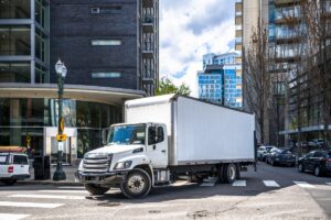 Big rig day cab white semi truck with long box trailer making local commercial delivery at urban city with multilevel residential apartments buildings turning on the city street with crossroad