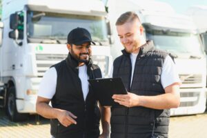 Two diverse truck drivers reviewing documents and smiling, standing outdoors next to parked semi trucks