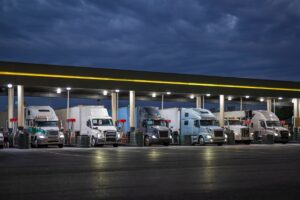 Professional big rig semi trucks with turned on headlights and loaded semi trailers standing on truck stop gas station at night time fill up the trucks tanks