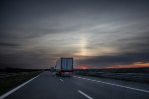 Semi-trucks driving on a highway at dusk with a cloudy sky and sunset in the distance
