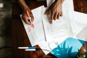 Hands reviewing and marking up financial documents with a red pen at a desk.
