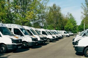 A row of white delivery vans and box trucks parked in a lot lined with trees, illustrating a small fleet ready for replacement planning