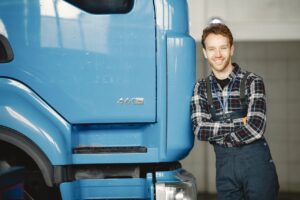 A smiling technician in a plaid shirt and overalls leans against the cab of a blue semi truck in a repair shop.