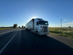 A white semi truck pulling a curtain-side trailer drives down a rural highway under a clear blue sky.