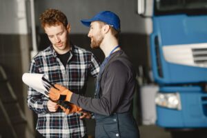Two technicians review a fleet maintenance checklist together in front of a blue semi truck in a service bay.