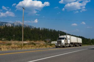 White semi truck with a trailer driving on a two-lane highway past forested mountains