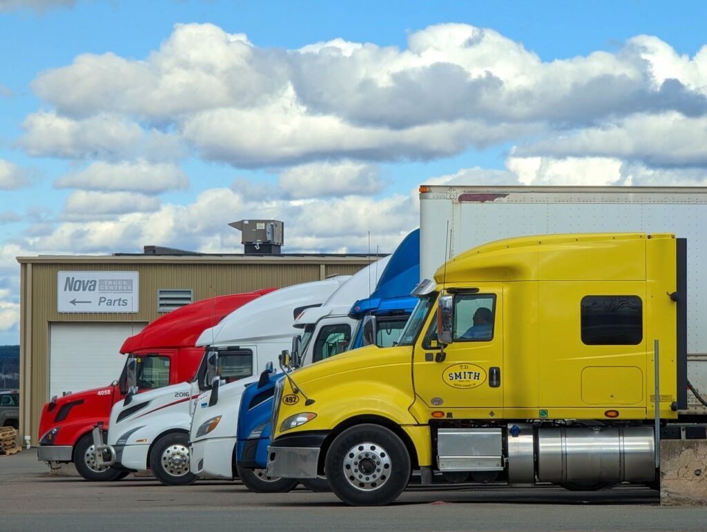 Line of red, white, blue and yellow semi truck cabs parked outside a truck parts building, showing heavy commercial vehicles that can count toward a year-end Section 179 semi truck purchase
