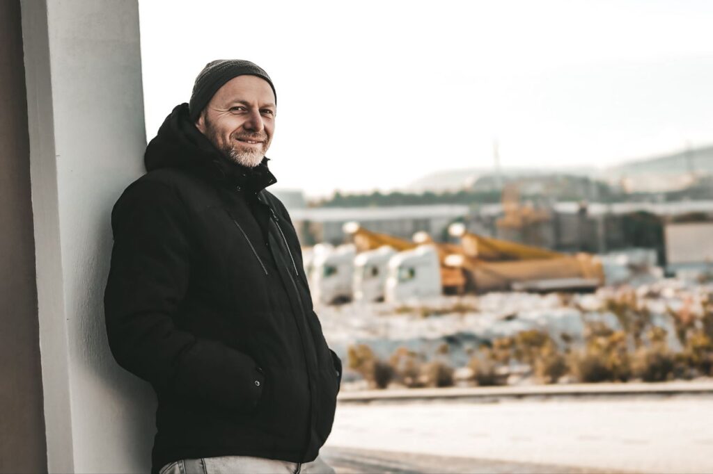 Smiling man in a beanie and black jacket leans against a wall near a lot of parked semi trucks