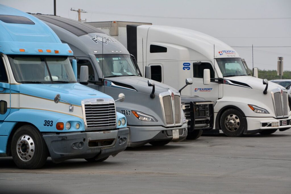 Three Class 8 semi trucks in light blue, gray, and white parked side by side in a truck lot