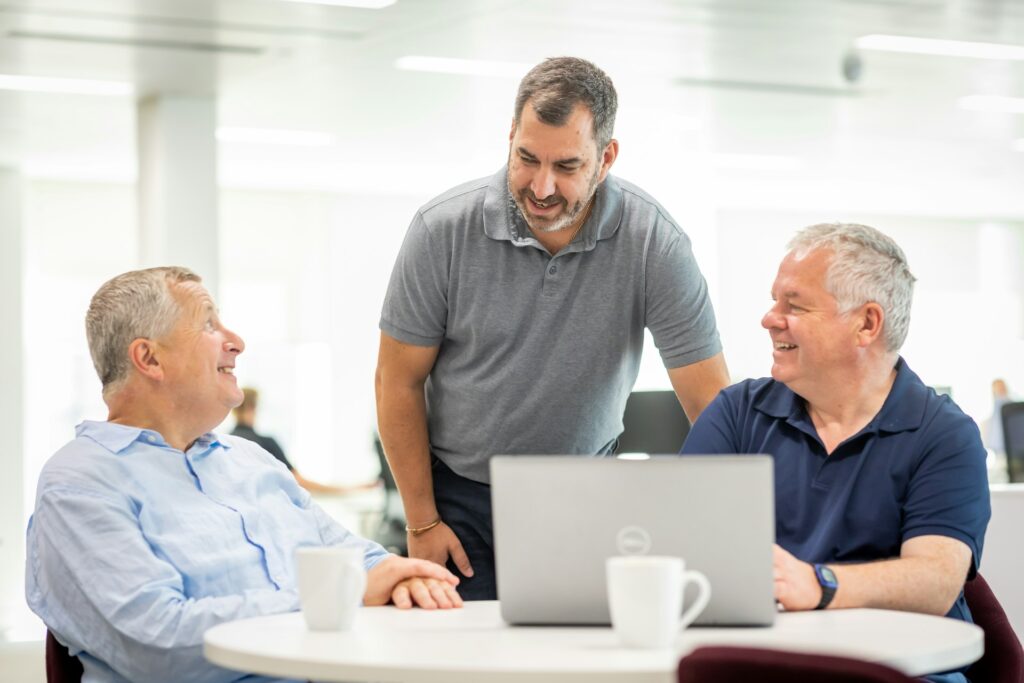 Three men talking and smiling around a laptop at an office table while reviewing truck loan options