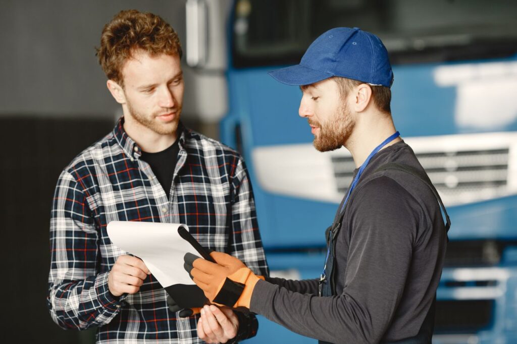Truck owner reviewing paperwork on a clipboard with a mechanic in front of a blue semi truck.