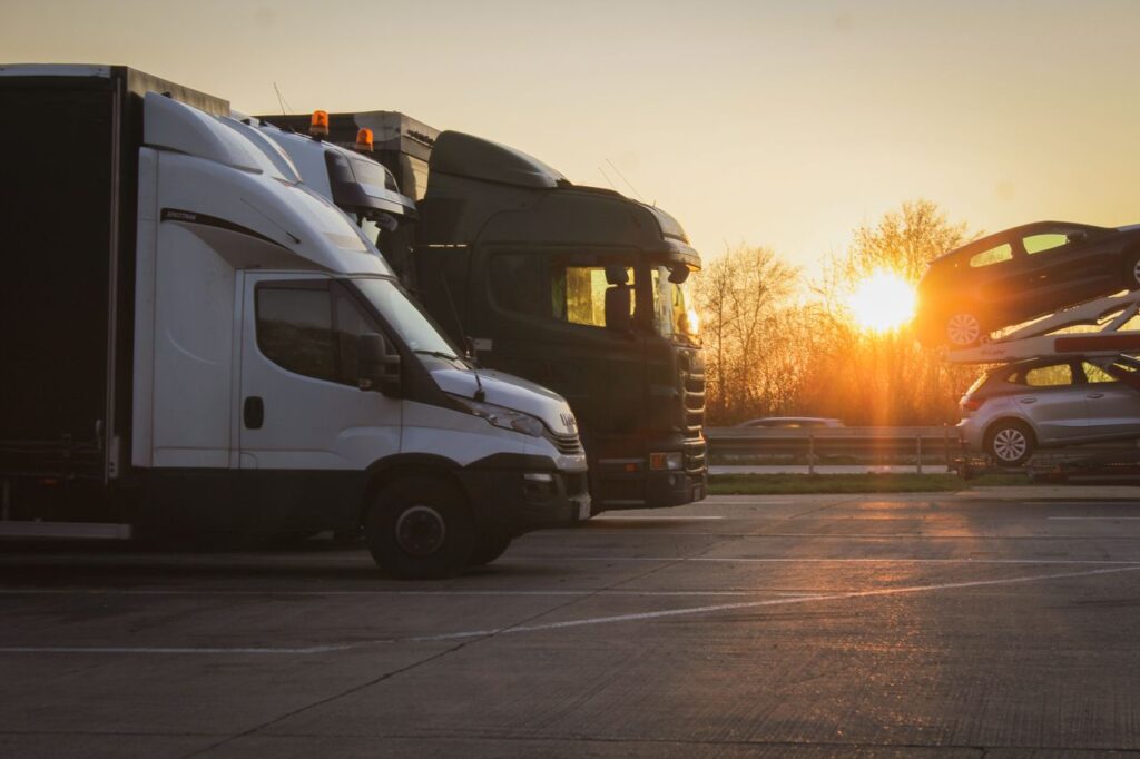 White box truck and dark semi truck parked at a truck stop at sunset, with a car carrier loaded with vehicles on the right