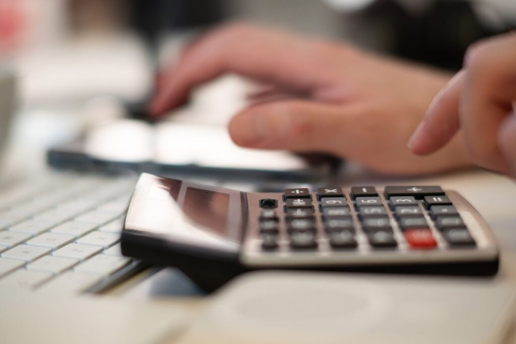 Close-up of a hand using a desk calculator next to a keyboard to work out monthly payments